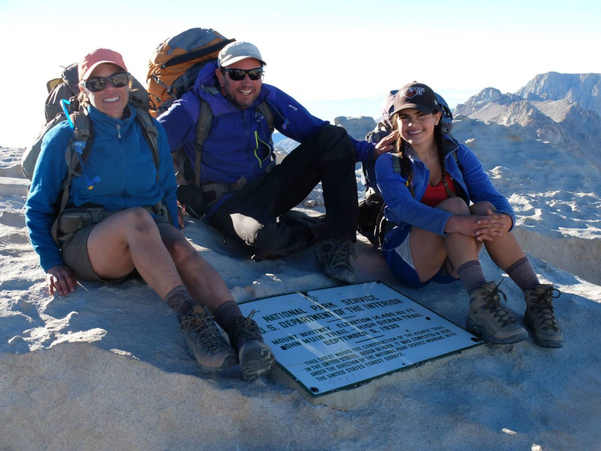 Debbie North and her husband and daughter smile for a photo on top of Mount Whitney with the National Park Service mountain marker between them